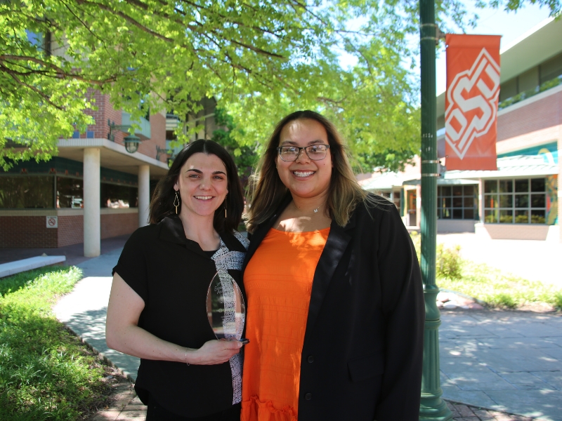 Two Health Promotion members stand smiling, holding a glass award, while standing in courtyard with trees and OSU-Tulsa in the background.