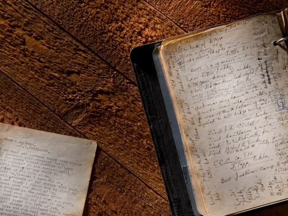 Old loose leaf page sitting next to old book with faded cursive text and discolored page margins on rough wood plank table