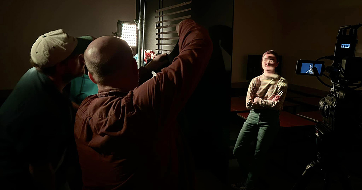Two people behind a camera work to light a female subject on a film set.
