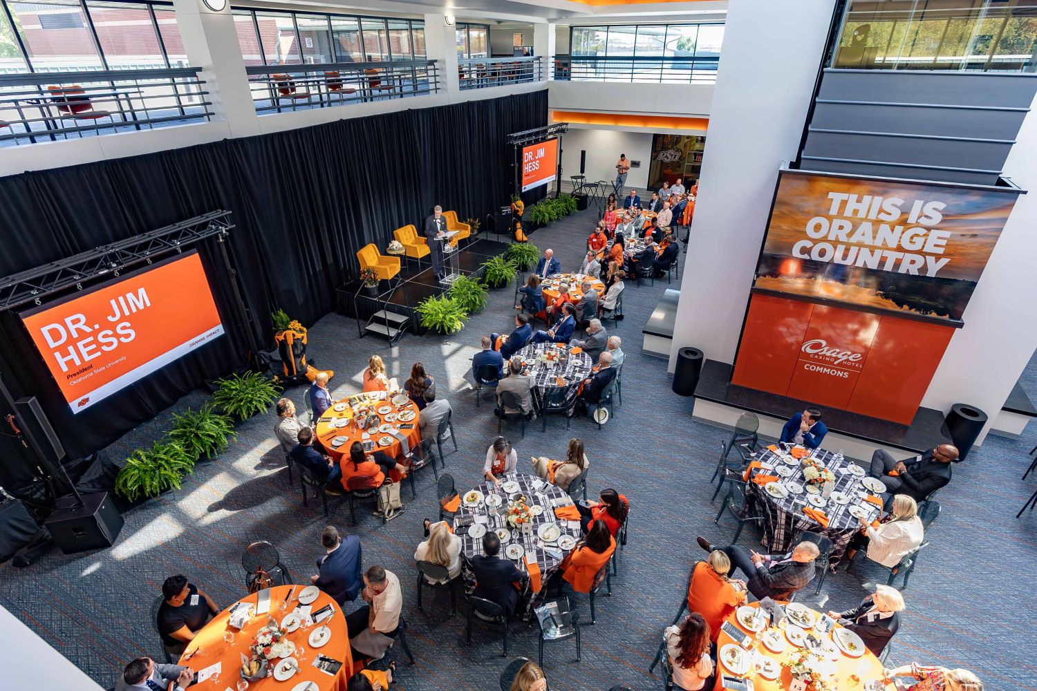 An overhead view of the Main Hall Commons, set for an event with multiple round tables and a stage with a screen that says "Dr. Jim Hess."
